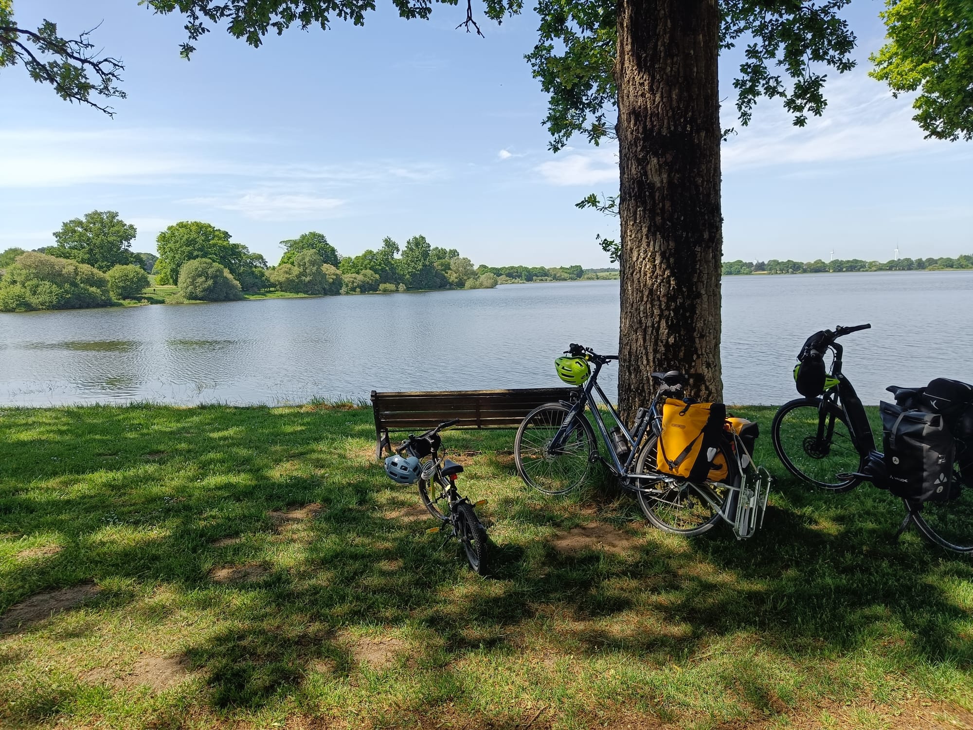 Photo de 3 vélos installés sur l'herbe, derrière un banc et devant un grand étang avec une vue sur la rive d'en face verdoyante. Le tout sous un grand ciel bleu.